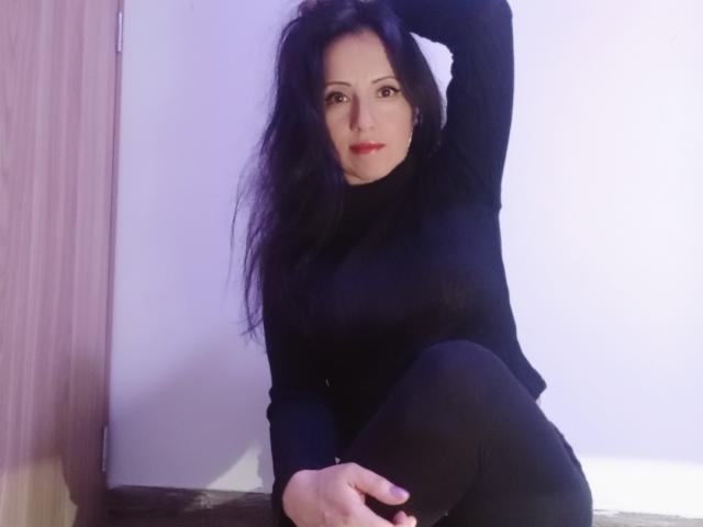 MondBlue has long black hair, wearing a black turtleneck and black pants, sitting with one arm behind her head and the other on her knee, showing her ass.