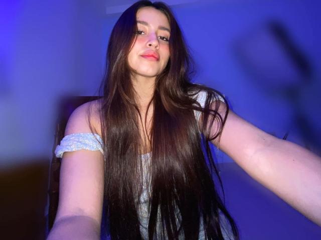 JulietaMorena with long brown hair, off-shoulder blue top, slight smile, blue light background, arms extended, holding camera. Boobs not visible, focus on face and hair.