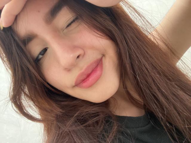 JulietaMorena with winking eye, dark brown hair, and pink lips, touching her head with a hand, wearing a black shirt, smiling subtly.