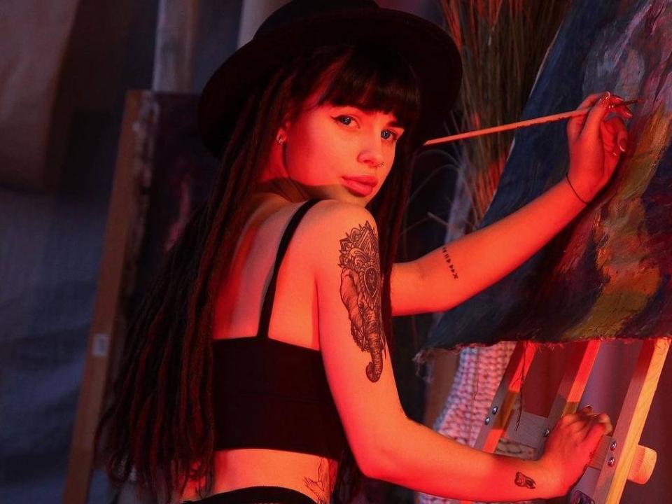 MaryJaness with long black dreads, black crop top, black hat, holding brush, painting, tattoo on right arm, red light, looking back, standing in front of canvas, holding a stick.