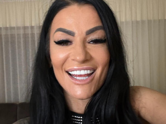 AndraD has dark hair, thick eyebrows, long black eyelashes, white teeth, and a shiny face. She's smiling with her mouth open, showing her teeth. She's wearing a black top.