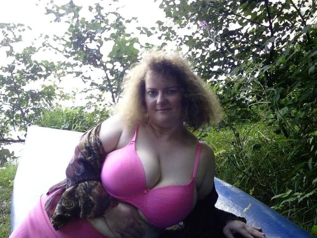 Vollbusige100dd-hot with curly blonde hair, pink bra showing big tits, and brown patterned shirt, sitting on blue surface, looking at camera, in green, leafy outdoor setting.