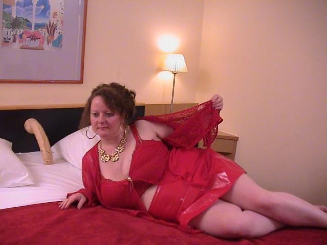 Vollbusige100dd-hot lying on a bed in red lingerie, holding up her sheer red skirt, exposing her large tits and red bra. She's wearing a gold necklace, looking slightly annoyed.