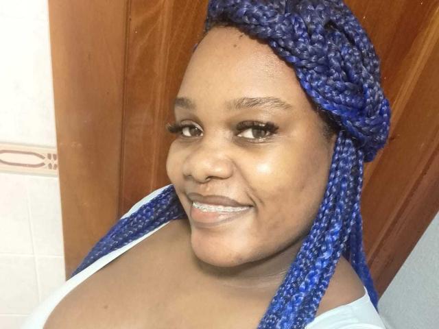 AgathaBlossom, con trenzas azules, sonriendo, mostrando sus pechos en una blusa blanca, ojos marrones y piel de color marrón claro, de pie frente a una puerta de madera..
