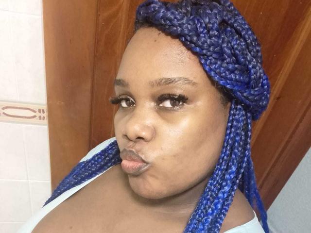 AgathaBlossom with blue braids, puffy lips, brown skin, wearing a white top, looking to the side, wooden door in background.