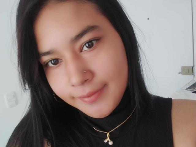 MeghanAss has smooth brown skin, black straight hair, and a small nose piercing. She's wearing a black sleeveless top and a gold necklace with a pearl. Smiling softly.