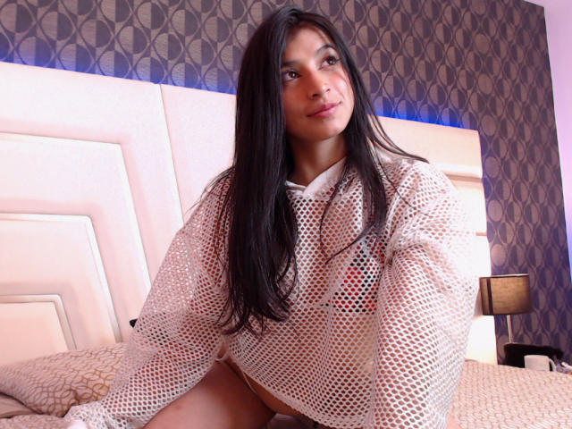 NaiaSanders with long black hair, wearing a white mesh top, sitting on a bed, showing her pussy through the holes, looking at the camera with a slight smile.