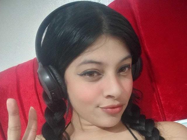 CamyHott69 with dark hair in braids, wearing black headphones, has light skin, brown eyes, and a slight smile, showing a finger gesture, red background.