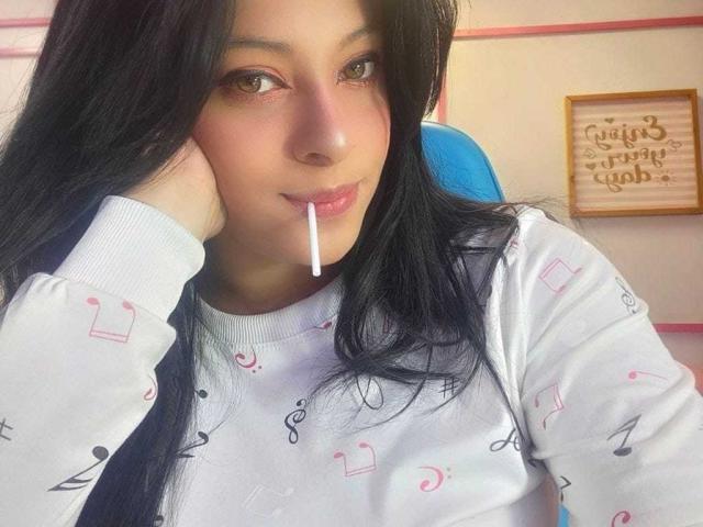 CamyHott69 hat langes, schwarzes Haar, braune Augen und trägt rosafarbenen Lippenstift, wobei sie eine Zigarette im Mund hat. Sie trägt ein weißes Hemd mit Noten und legt ihren Kopf auf ihre Hand..