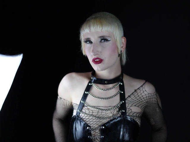 Lynn-Tonic-hot with short blonde hair, red lipstick, black leather harness, fishnet sleeves, and metal chains, looking fierce with exposed tits and a punk rock vibe.