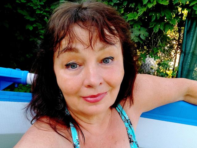 MilaMils has medium brown hair, blue eyes, and is smiling with a slight pink lip. She's wearing a blue patterned halter top, sitting in a blue and white boat.