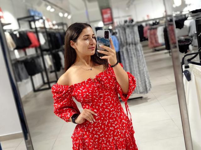 Mirellia, con un busto de tamaño medio, vestida con un vestido rojo con estampado floral de corte asimétrico, se está tomando un selfie frente a un espejo en una tienda, sosteniendo un teléfono con una funda azul. Tiene el pelo largo y castaño, y una expresión tranquila..