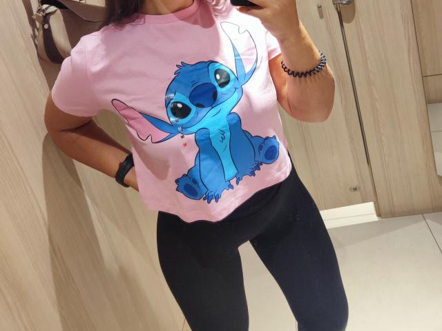 Mirellias rosafarbenes Hemd mit einem blauen Stitch-Motiv, schwarze Leggings und ein schwarzes Armband, stehend in einer Umkleidekabine, ihre Hand auf der Hüfte, eine Kamera in der anderen Hand..