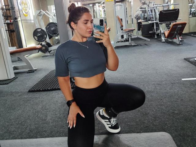Mirellia with her tits in a tight gray crop top, black leggings, and black sneakers, squatting in a gym, holding a phone, wearing a black watch.