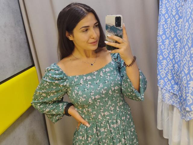 Mirellia, con cabello castaño oscuro, un vestido floral verde, mangas abullonadas, con una mano en la cadera, tomando una selfie en el espejo, sonriendo, llevando un collar y una pulsera, sosteniendo un teléfono, en un probador..