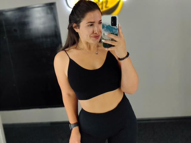 Mirellia with dark hair in a ponytail, wearing a black sports bra and high-waisted black pants, taking a mirror selfie, showing her tits and ass, smiling slightly.