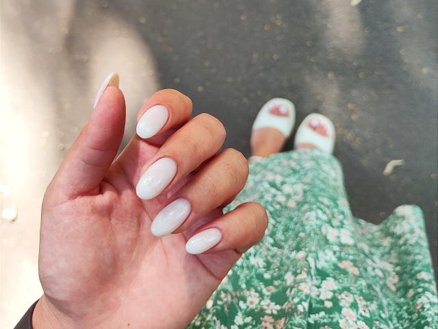 La mano de Mirellia, con uñas largas, blancas y en forma de almendra. Lleva un vestido verde con estampado floral y zapatos blancos de tacón abierto, que se ven en la parte inferior. Su piel es clara y suave..