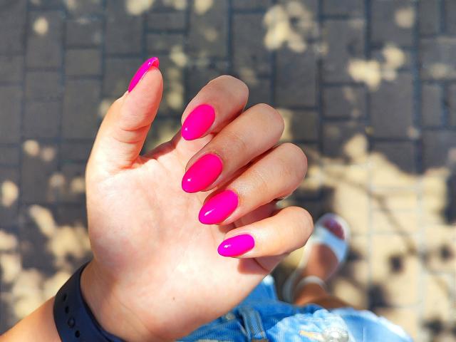 La mano de Mirellia, con uñas de color rosa brillante y forma almendrada, que muestran sus uñas brillantes y lustrosas. Lleva una pulsera negra y pantalones vaqueros azules, y en el fondo se ve una sandalia blanca..