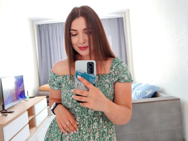 Mirellia mit langen, braunen Haaren, einem grünen, blumigen Kleid, das von einer Schulter abrutscht, macht ein Selfie und zeigt dabei leicht ihre Brüste, lächelt, trägt rosafarbenen Lippenstift, hält ein blaues Telefon und hat eine weiße Wand im Hintergrund..