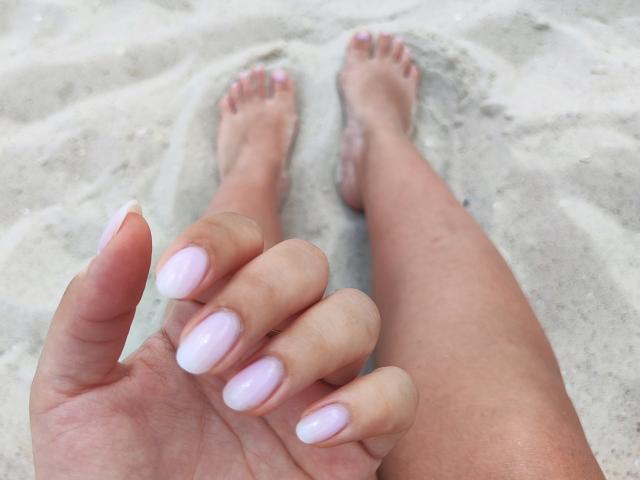Mirellia's hand with pink nail polish, fingers slightly spread, resting on sandy beach, legs in background, toes partially covered in sand, close-up shot.