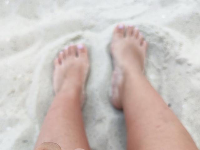 Mirellias Zehen sind mit Sand bedeckt, ihre Beine stehen leicht auseinander, wodurch ihre rosa lackierten Zehennägel sichtbar sind, und sie trägt ein kleines Armband an ihrem linken Knöchel..