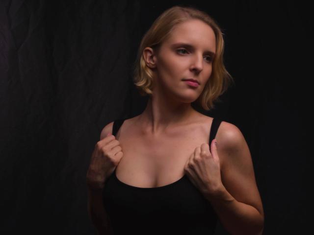 Luna-Lacroix-hot with blonde bob, holding black tank top straps, exposing her small tits, standing against dark background, looking slightly to the side with a serious expression.