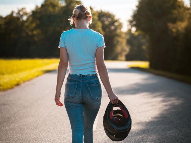 Luna-Lacroix-hot, white tee, blue jeans, holding black helmet in right hand, standing on empty road, back view, sunlit, forested background, blonde hair in bun, afternoon light.