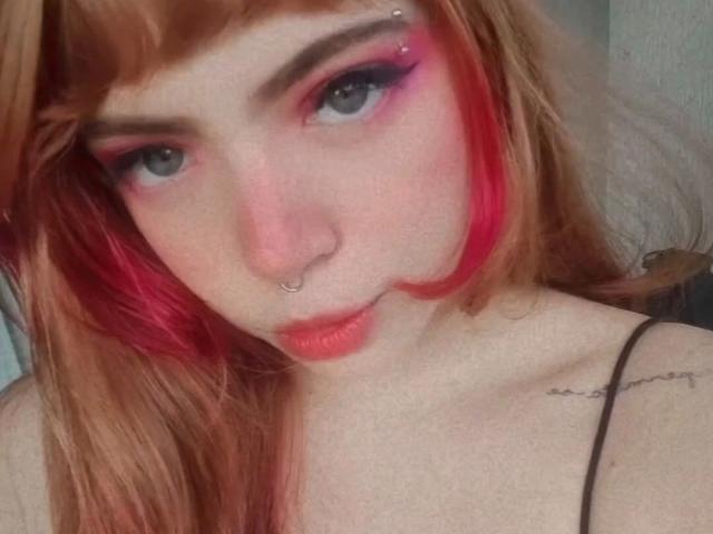 Acarolleao-hot with red hair, pink eye makeup, and nose ring, sucks on a black candy, wearing a black top, sitting on a bed with blue wall and string lights.