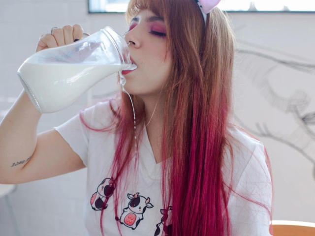 Acarolleao-hot has pink and red hair, a nose piercing, and black eyeliner with pink eyeshadow. She's looking down with pouty red lips and a tattoo on her shoulder.