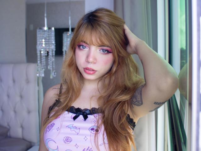 Acarolleao-hot with red hair, wearing a pink halter top, blue jeans, and sunglasses on head, showing a tattoo on right arm, standing outdoors, hands in hair.