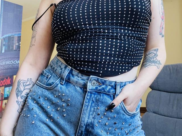 KarKutie has small, pierced tits in a black, studded crop top and high-waisted, blue, studded jeans. She's tattooed with a hand in her pocket.