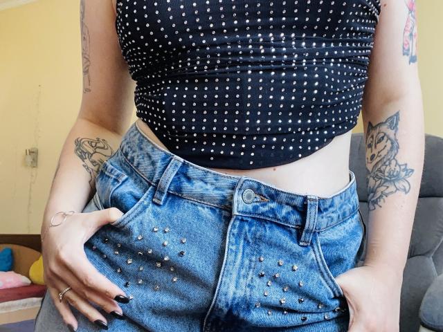 KarKutie's black, polka-dot crop top and blue, studded, high-waisted jeans. She's got tattoos on both arms and black nail polish, with hands in her jeans.