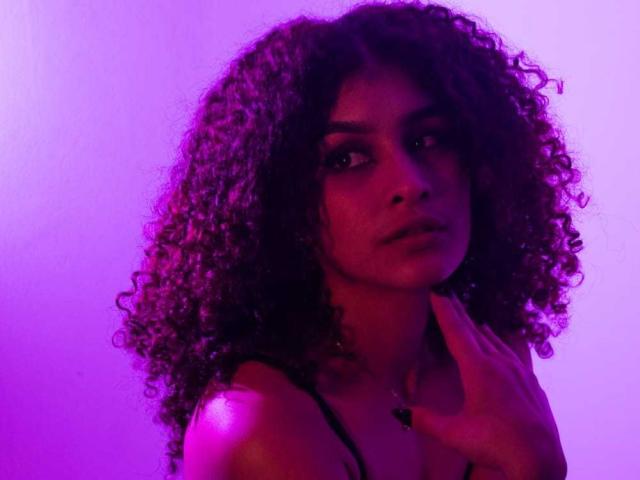 Amaia Escotet, com cabelos escuros e cacheados, iluminação em tons de roxo e rosa, olhando de lado, com a mão perto do pescoço, vestindo uma blusa preta com alças, com uma expressão sensual e séria..