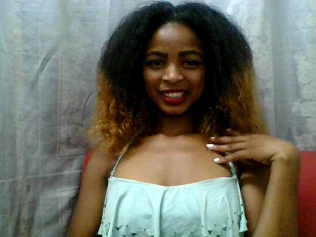 AlexaChan with curly black and blonde ombre hair, smiling, wearing a light blue strapless top, showing small tits, red lipstick, and white nails, hand on chest.