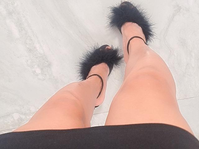 Die Beine von NaughtySecrets in einem schwarzen Rock, mit schwarzen, pelzigen High Heels mit Riemchen um den Knöchel, aufgenommen aus einem niedrigen Winkel. Ihre Beine sind hellhäutig und leicht glänzend..