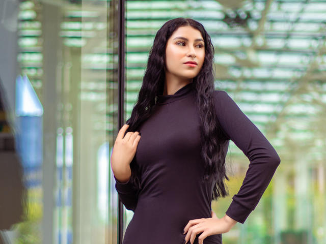 EmaHenderson with long black hair, wearing a tight black long-sleeve dress, standing near a glass wall, hand on chest, looking confident, with a slight smirk on her face.