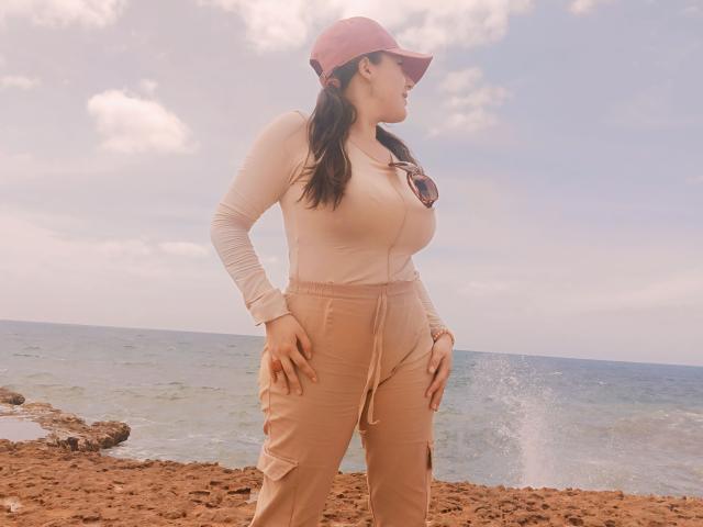 MabyLegrand with big boobs, tan pants, pink long-sleeve top, pink cap, and pigtails stands on rocky shore, hands on hips, ocean and cloudy sky in background.