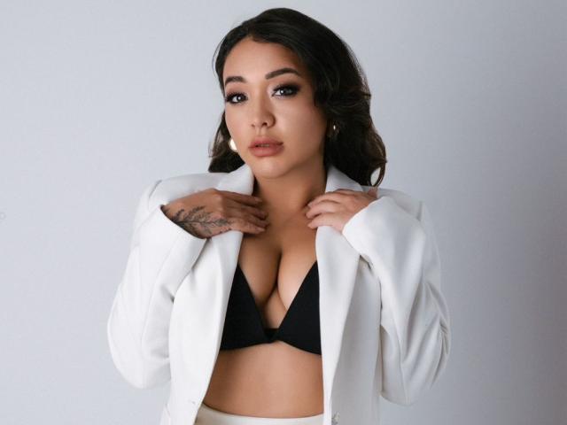 MiaWheel with dark wavy hair, wearing a white blazer over a black bra, hands on jacket, tattoos on right hand, subtle makeup, looking at camera, neutral expression.