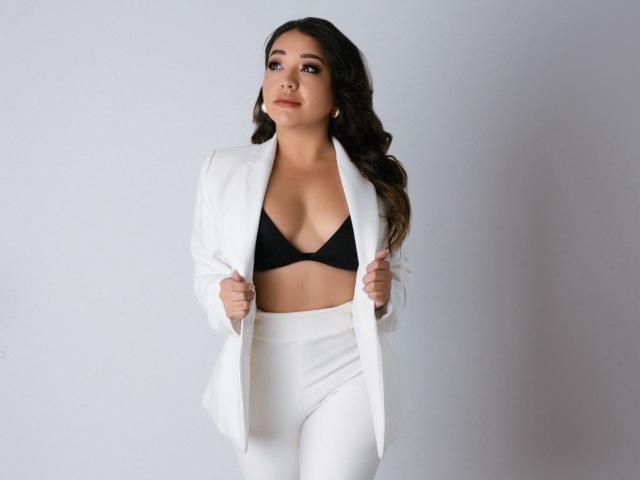 MiaWheel with dark wavy hair, wearing a white blazer, black bra, and white high-waisted pants, looking slightly to the side, hands on blazer. Boobs visible.