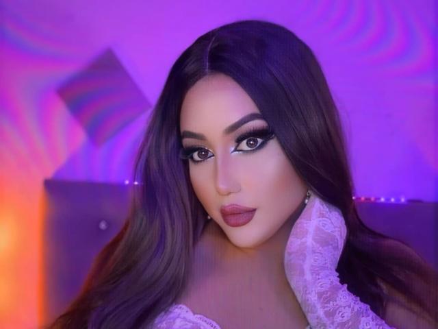 AntonellaMistresss has long brown hair, dark eyeliner, and full pink lips. She's wearing a white lace collar and looking directly at the camera with a seductive expression.