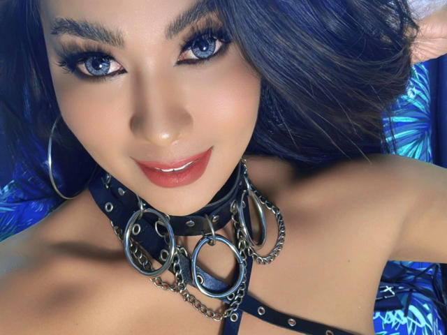 AddictiveMiss4u-hot with blue eyes, dark hair, and red lips, wearing a black leather collar with metal rings, and large silver hoop earrings, smiling at the camera.