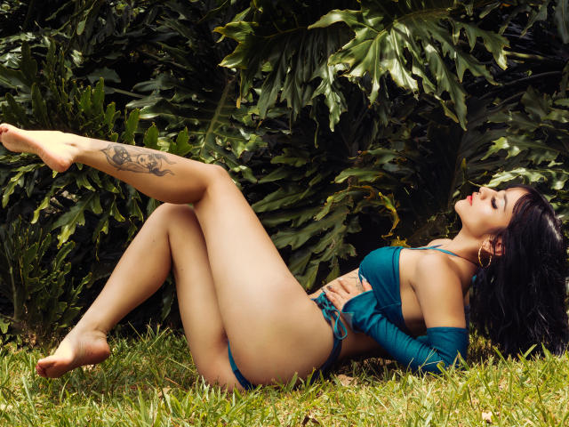 LindaWatsong is laying on grass with legs up, wearing a green bikini, showing her tits and ass, tattoo on her right thigh, black hair, red lipstick, and green leaves.