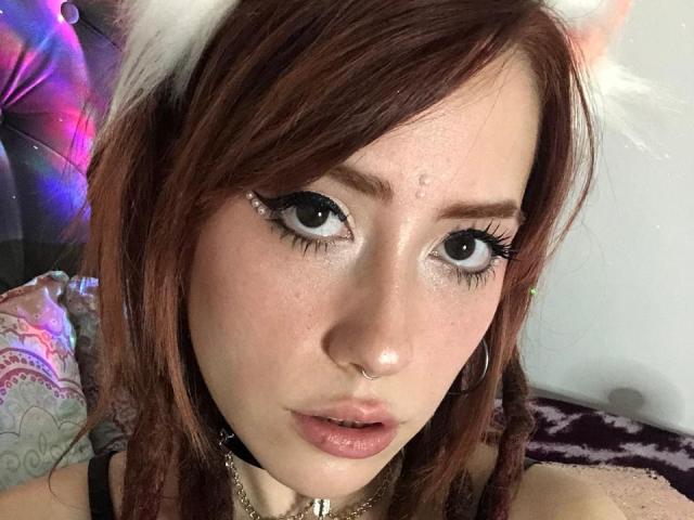 SarahDav mit Katzenohren, braunen Haaren, großen blauen Augen, glitzerndem Eyeliner, einem Nasenring, Creolen, einer Halskette und rosafarbenen Lippen. Außerdem ist ein leichter Glanz auf ihrem Gesicht zu sehen. Ihre Brüste sind sichtbar..