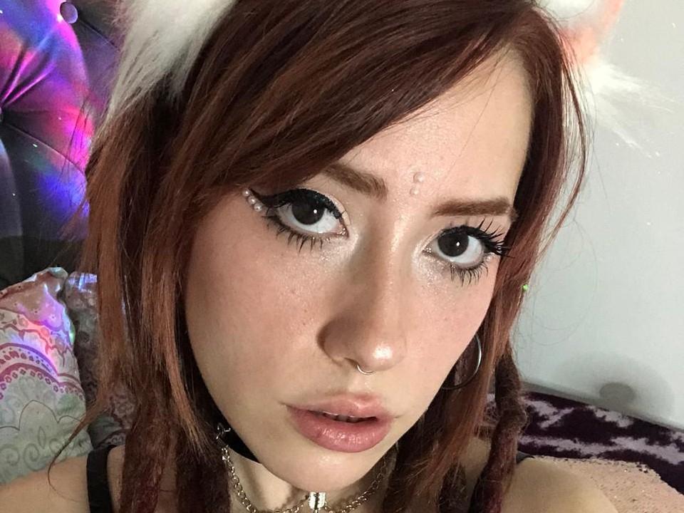 SarahDav mit Katzenohren, braunen Haaren, großen blauen Augen, glitzerndem Eyeliner, einem Nasenring, Creolen, einer Halskette und rosafarbenen Lippen. Außerdem ist ein leichter Glanz auf ihrem Gesicht zu sehen. Ihre Brüste sind sichtbar..