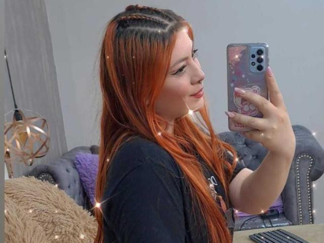 IsabellaRobertz met lang rood-oranje haar, een zwart shirt, die een selfie maakt in een spiegel, een roze telefoonhoesje met een sterrenstelsel patroon vasthoudend, lachend met een schattig gezicht, in een gezellige kamer..