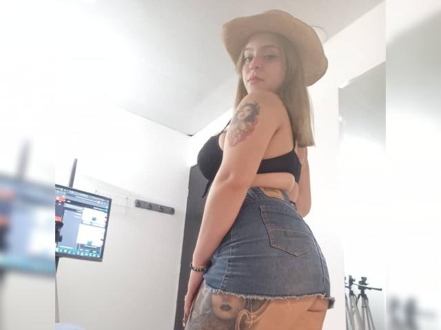 StacyAnzola, avec un chapeau marron, un soutien-gorge noir et une jupe en jean, montrant un tatouage sur le bras droit, les fesses apparentes à travers la jupe déchirée, regardant en arrière, debout dans une pièce lumineuse, appareil photo à gauche..