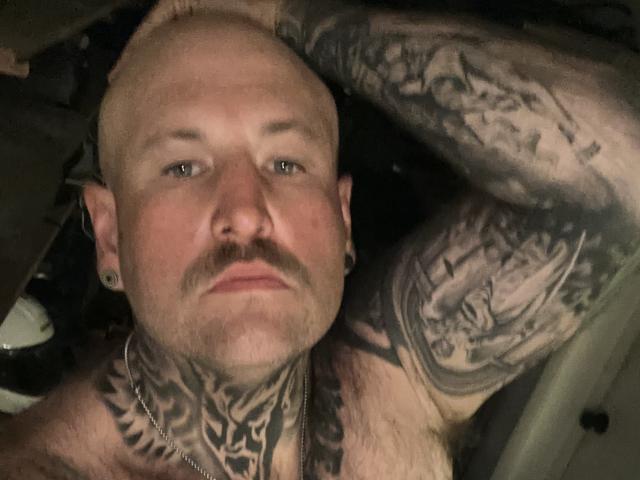 TattedDody is een kale man met een snor en tatoeages op zijn nek, borst en arm, die een ketting draagt en oorbelletjes heeft, en die ligt te rusten met een serieuze blik op zijn gezicht..