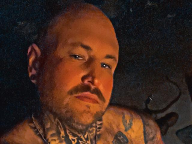 TattedDody è un uomo calvo, tatuato e con i baffi, che guarda di lato con un'espressione seria, indossa una collana a catena, in una foto scura e con una texture particolare..
