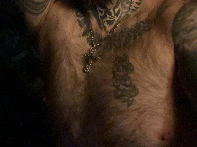 De harige borst van TattedDody, met tatoeages, waaronder een kruis en ingewikkelde ontwerpen, is zichtbaar in gedimd licht. Zonder borsten, gefocust op het torso. Donkere, schaduwrijke achtergrond..