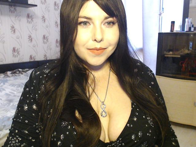 WhitteLotus with long brown hair, fair skin, and orange lipstick, wearing a black floral top with a deep V, showing her big boobs and a silver necklace.